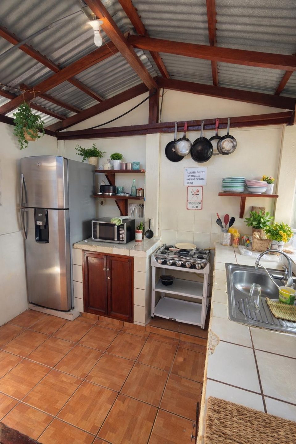 Shared kitchen at Hostal Los Chamos — fridge, gas stove, hanging pans and open shelving under a corrugated roof
